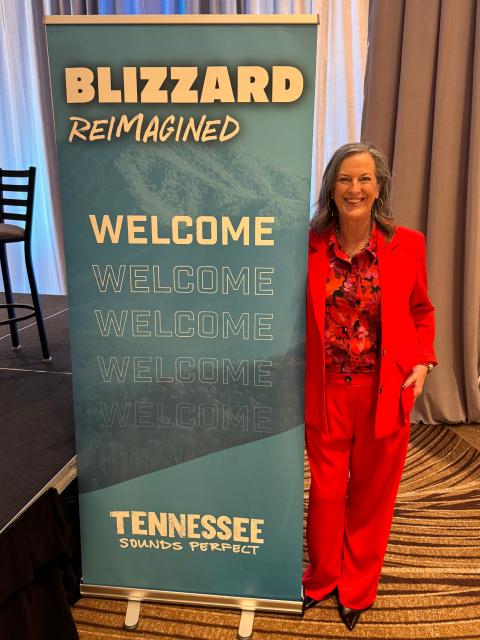Person standing indoors beside a vertical event banner that reads “Blizzard Reimagined” and “Welcome,” with “Tennessee Sounds Perfect” at the bottom, on a conference stage with curtains and patterned carpet visible.