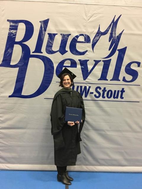 A graduate in cap and gown holds a diploma while standing in front of a large “Blue Devils UW–Stout” backdrop.