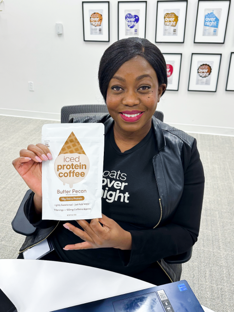 Person seated at a table holds a package labeled “Iced Protein Coffee, Butter Pecan,” with framed product posters displayed on the wall behind.