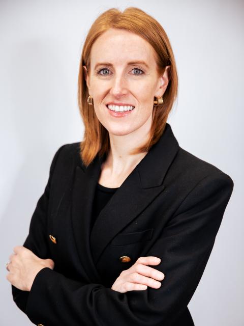 Person wearing a black blazer with gold buttons stands with arms crossed against a light gray background.