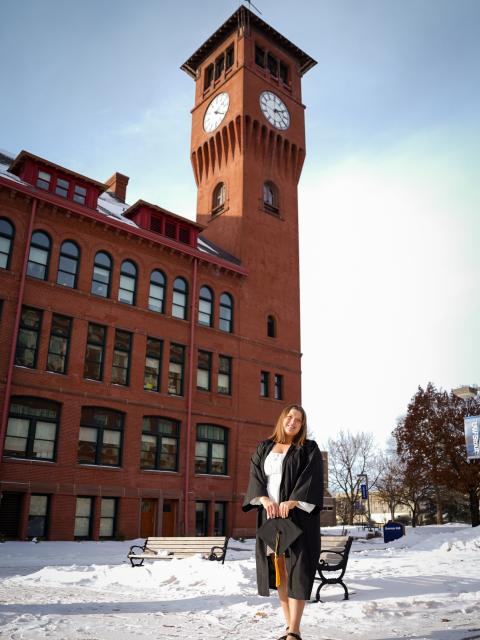 UW‑Stout graduate in cap and gown stands outdoors in front of Bowman Hall and the campus clock tower on a winter day.