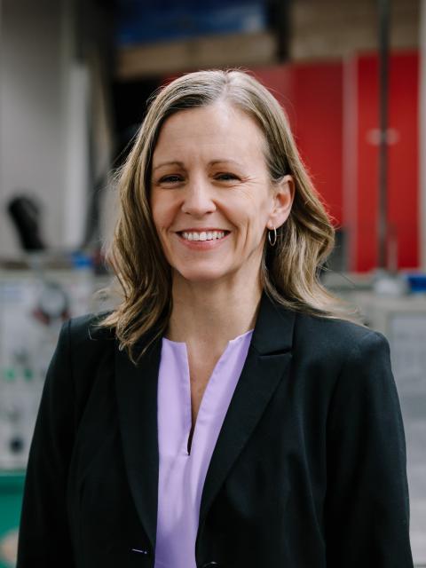Person in a black blazer and light purple blouse, smiling at the camera, standing in an indoor lab or workspace with blurred equipment and red storage cabinets in the background.