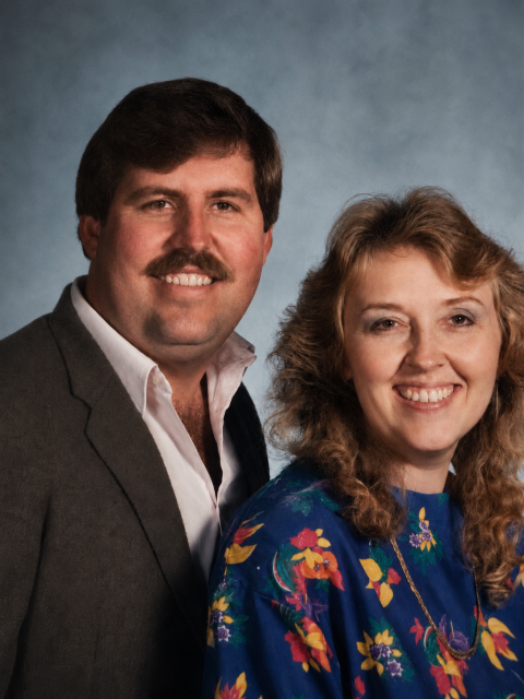 Studio portrait of two adults posed side by side against a neutral blue background, both facing the camera.