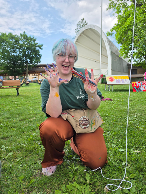 Person kneels on grass holding paintbrushes and displaying paint-covered hands in front of an outdoor mural project.