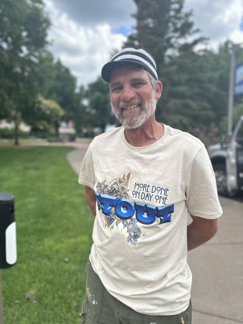 Portrait of a mural artist standing outdoors near a sidewalk and grassy area wearing a paint-splattered T-shirt.