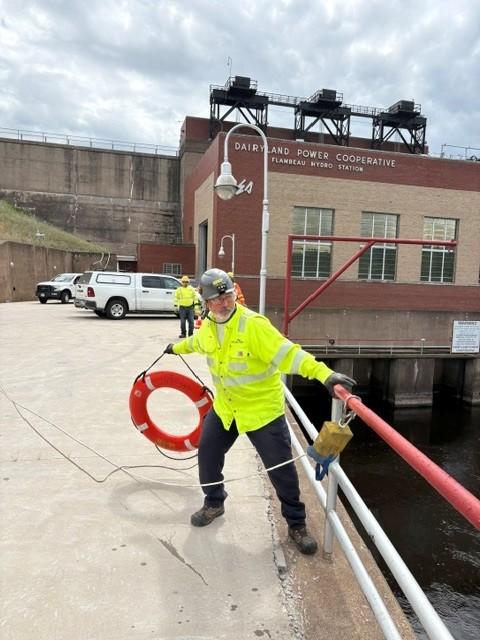A utility worker wearing safety gear demonstrates a water rescue exercise near a hydroelectric facility while holding a life ring.