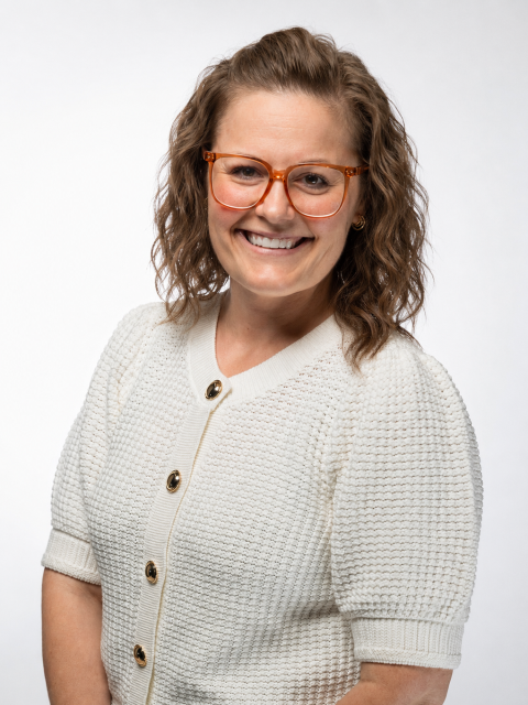 Professional studio headshot of a person with shoulder-length wavy hair and large amber-colored eyeglasses, wearing a textured white short-sleeve cardigan with gold buttons, posed against a light gray background.
