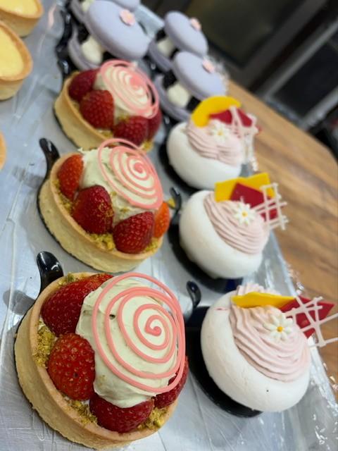 An assortment of decorated pastries displayed on a tray, including strawberry tarts topped with cream and pink spiral decorations, alongside white dome-shaped desserts with pink frosting accents.