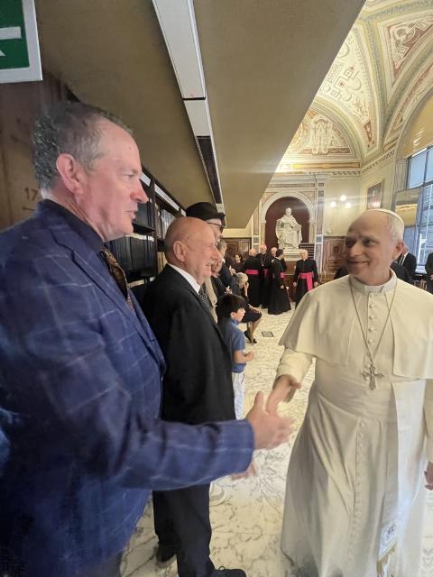 A person in a blue jacket greets a religious leader dressed in white inside an ornate marble hall with vaulted ceilings and statues in the background.