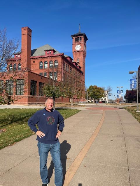 Poul David “Dave” Rasmussen stands on a campus walkway in front of UW-Stout Polytechnic's Bowman Hall and clock tower on a sunny fall day.